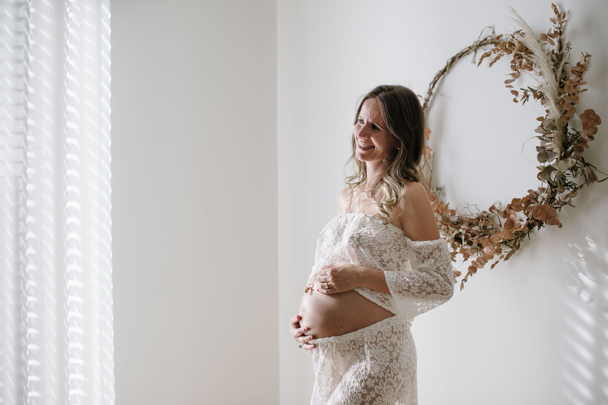 Side view maternity photo with mother in lace dress against white wall and window light.