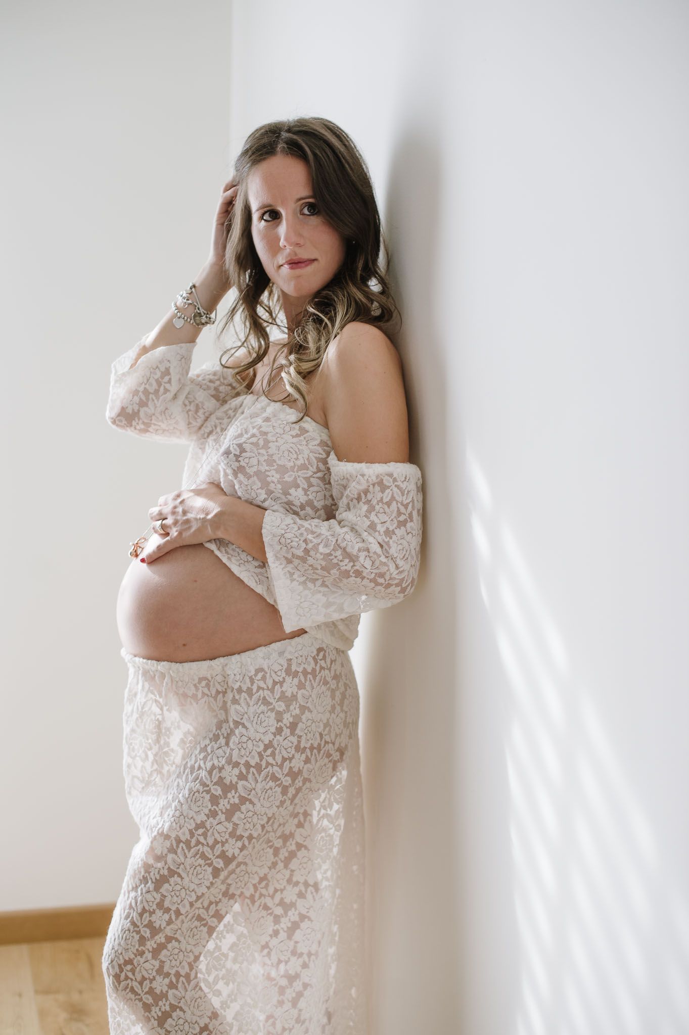 Full length maternity portrait of an expectant mother in white lace dress against a wall.