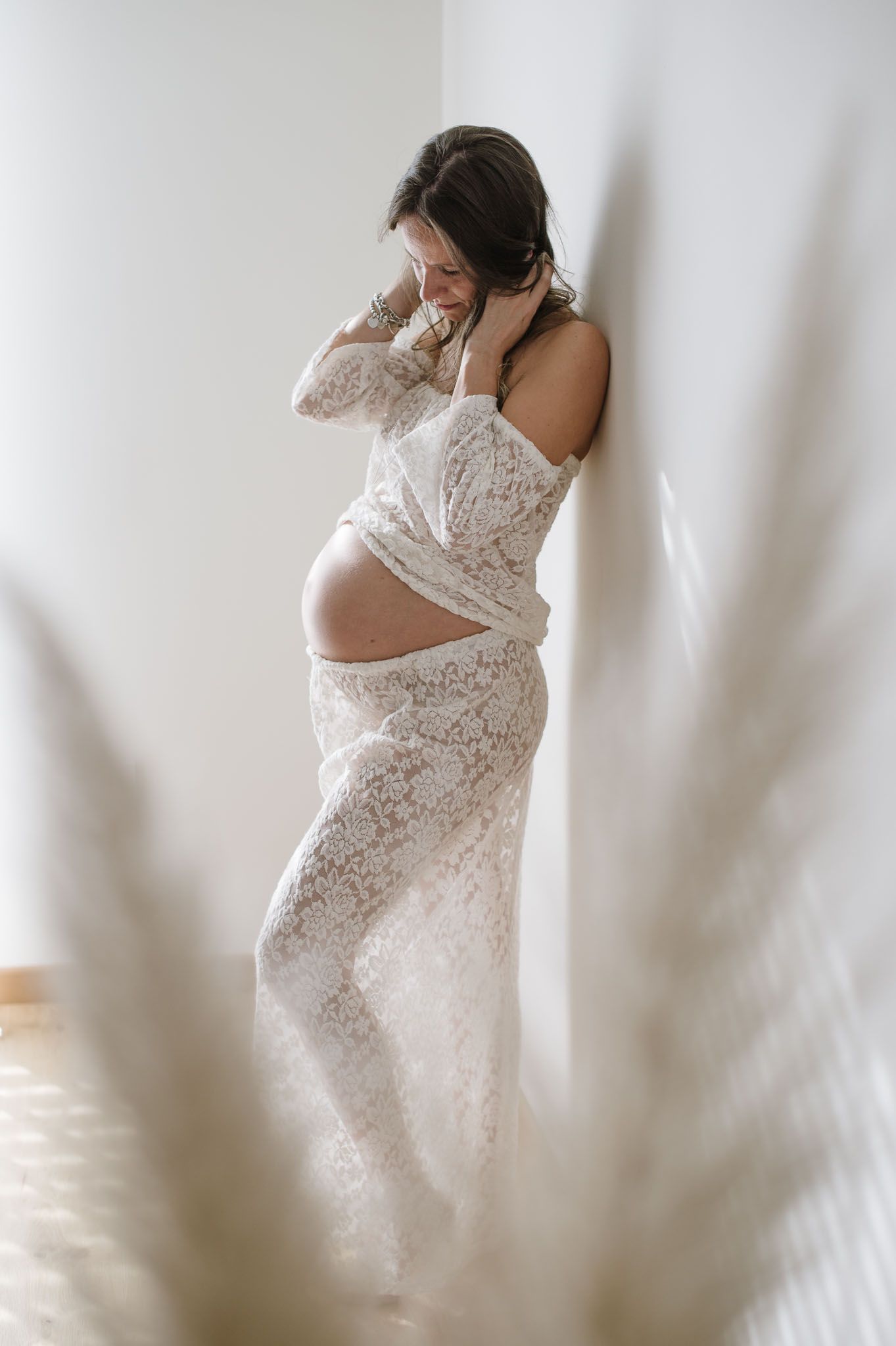 Artistic maternity photo with soft foreground blur showing mother in white lace dress.