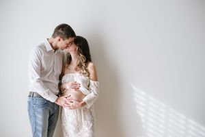 Tender maternity portrait of couple sharing intimate moment against white wall with window light.