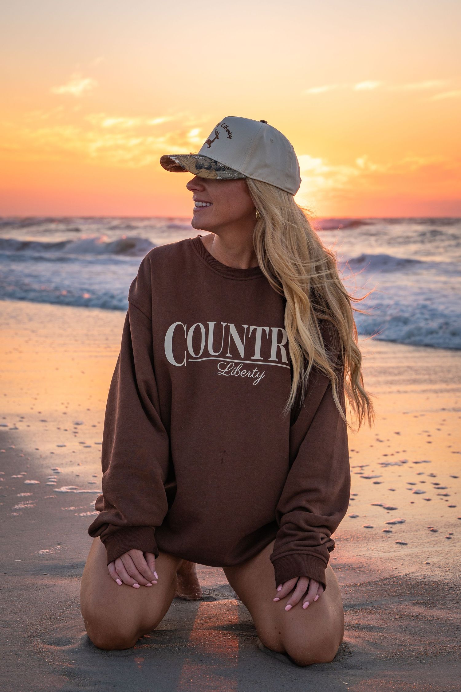 Sunset beach scene with lifestyle fashion elements including brown sweatshirt and baseball cap.