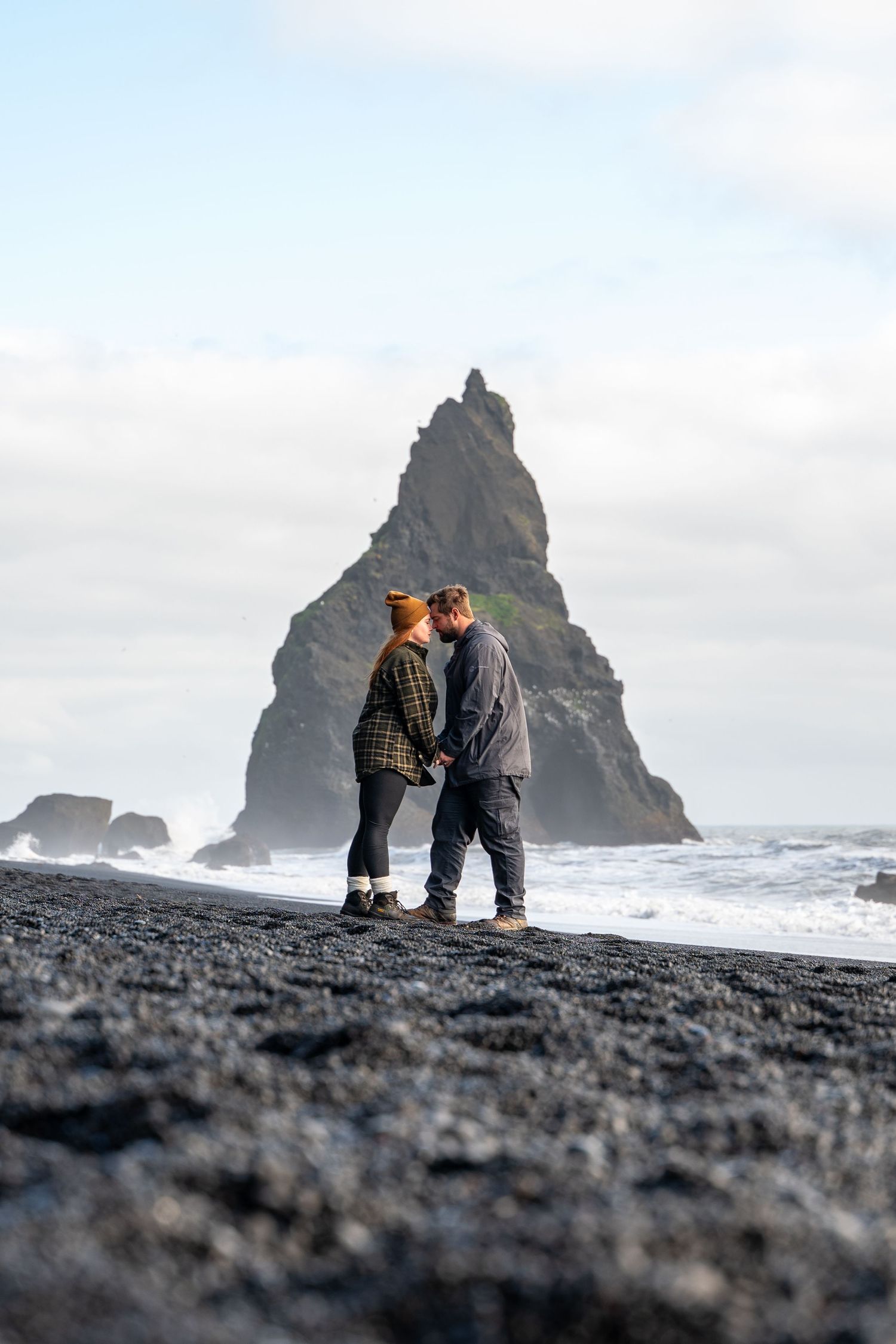 Couple sharing intimate moment on black sand beach with dramatic rock formation in Iceland.