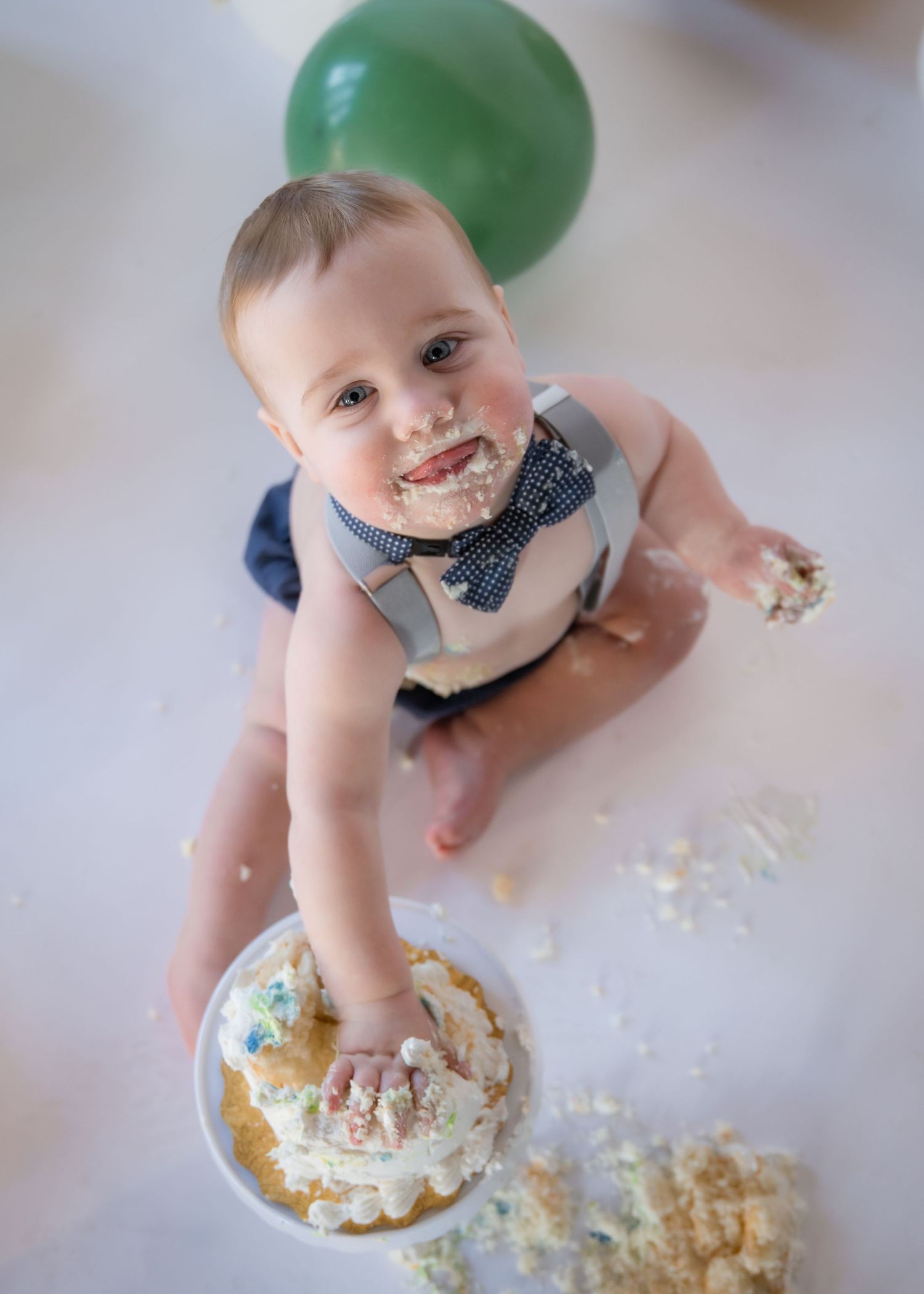 A joyful baby wearing a bow tie and suspenders sits on the floor with a smashed birthday cake and green balloon nearby.
