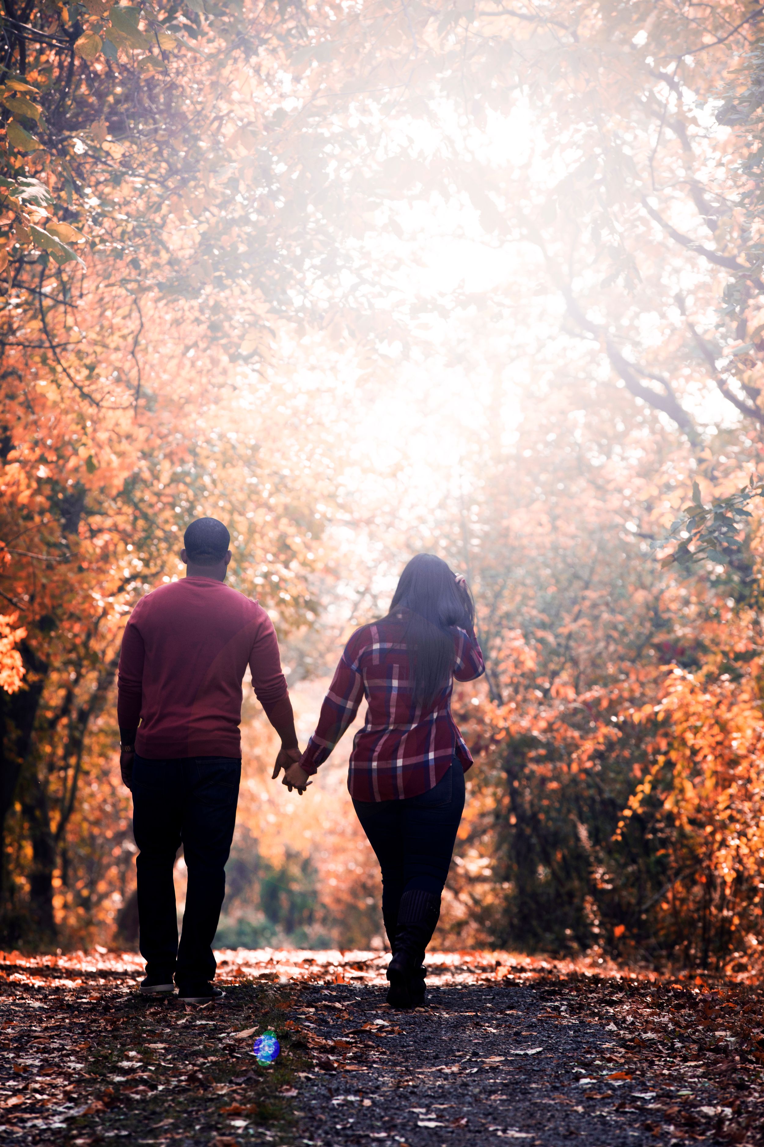A couple holding hands while walking on a leaf-covered path surrounded by autumn foliage and golden sunlight.