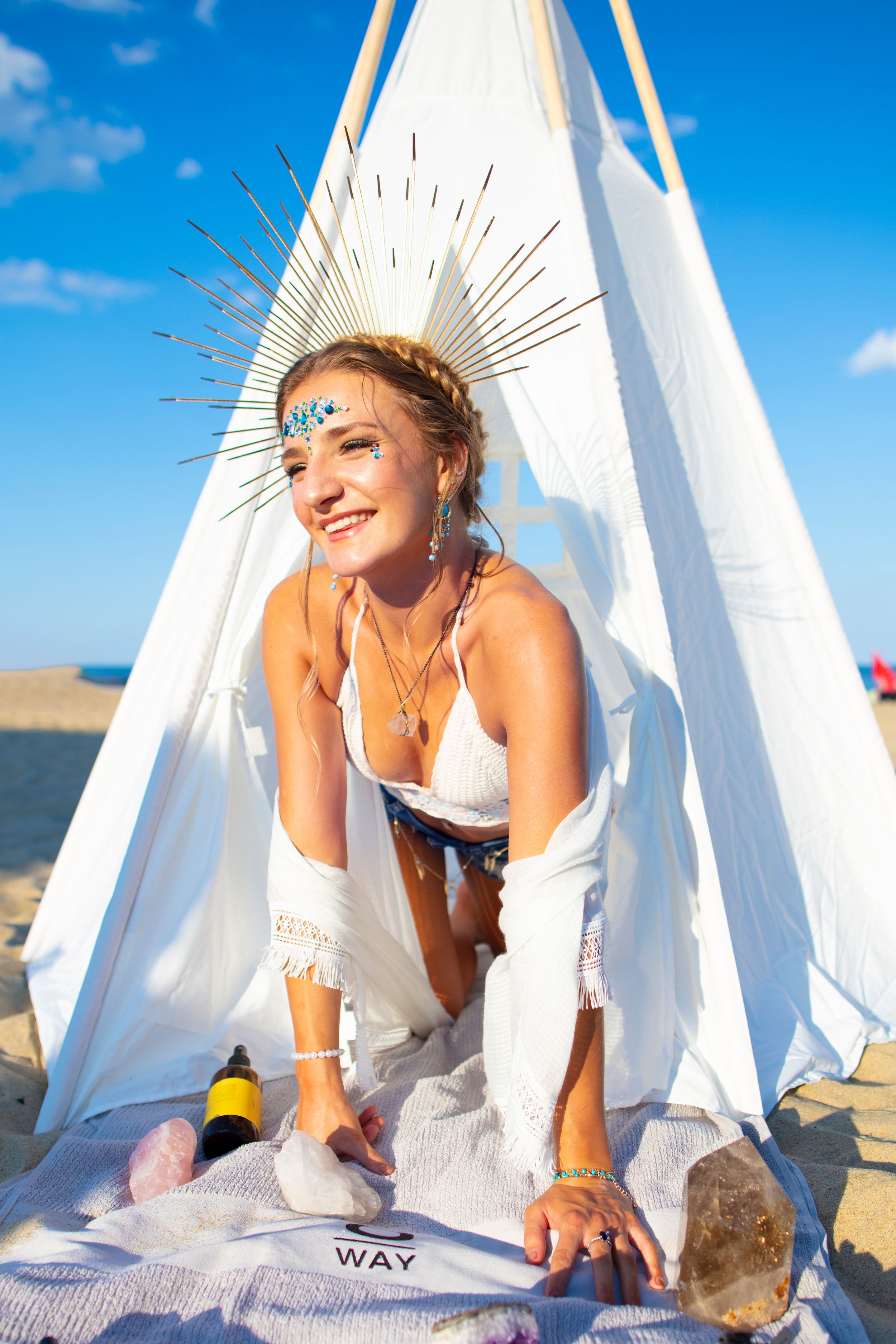 A woman in festival attire and face jewels poses on a white teepee tent at a beach during a sunny day.