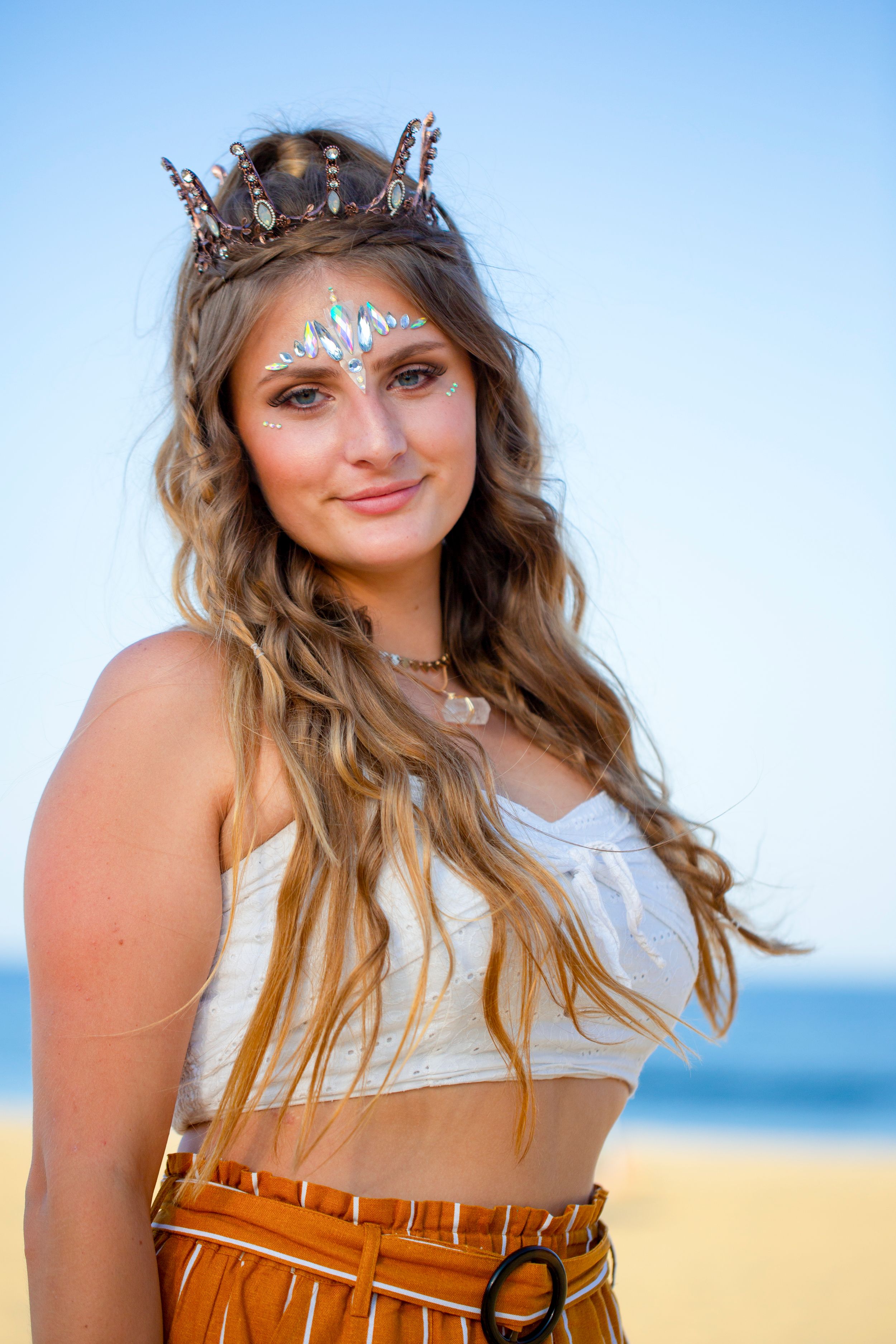 A person wearing a crown and face jewels poses on the beach in a white crop top and brown skirt.