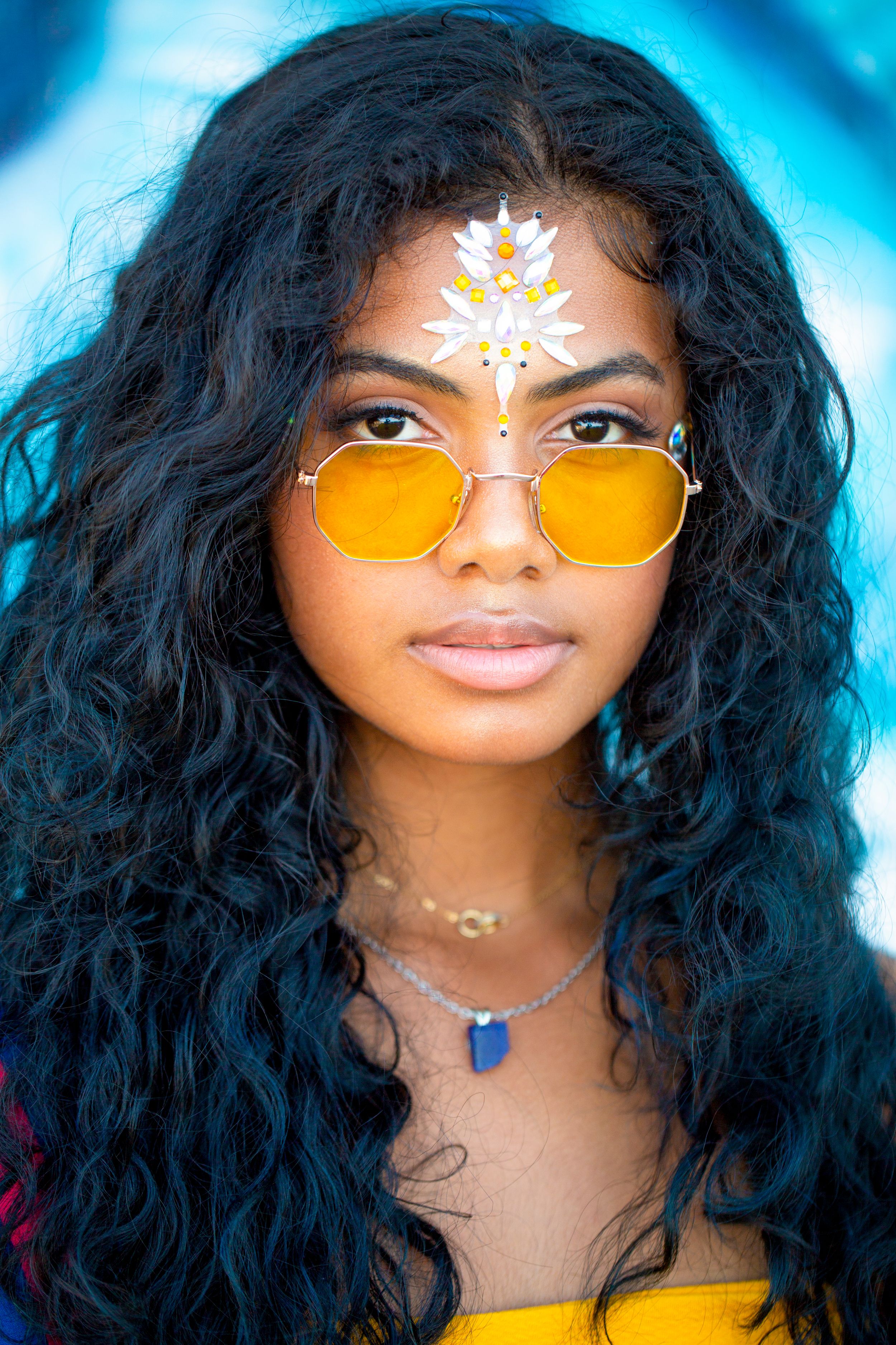 Portrait showing yellow-tinted sunglasses and intricate face jewels with long wavy hair.