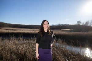 A person stands near a marsh at sunset with tall grasses and a serene waterway in the background.