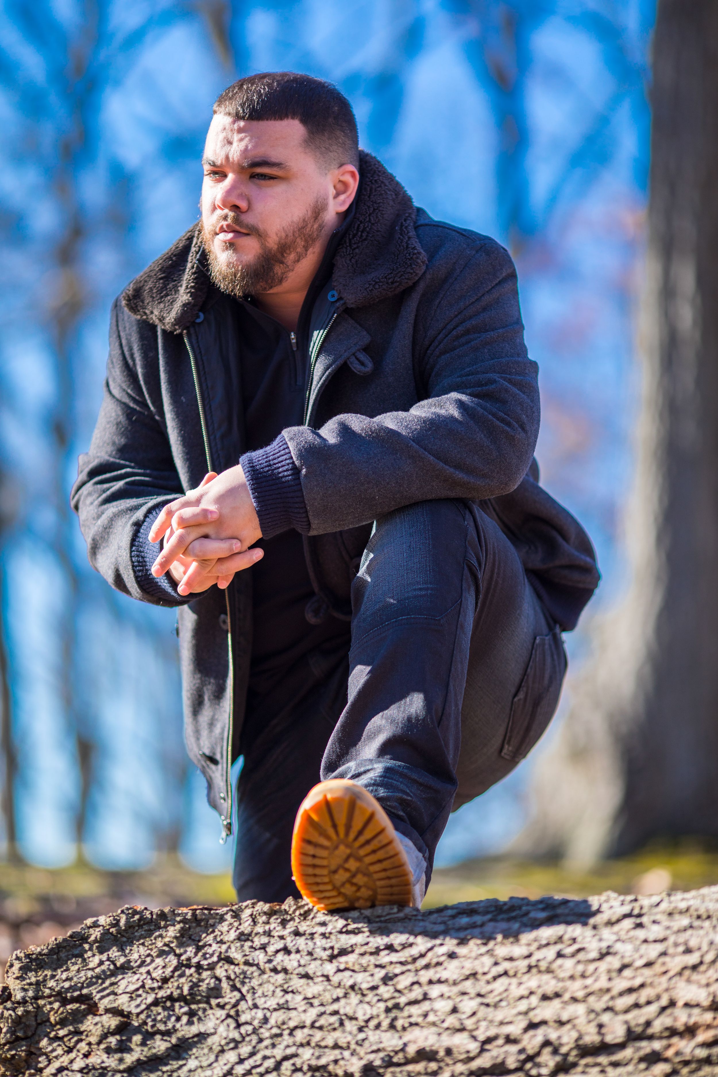 A person in a black winter coat crouches on a rock outdoors against a blue sky and trees on a sunny day.
