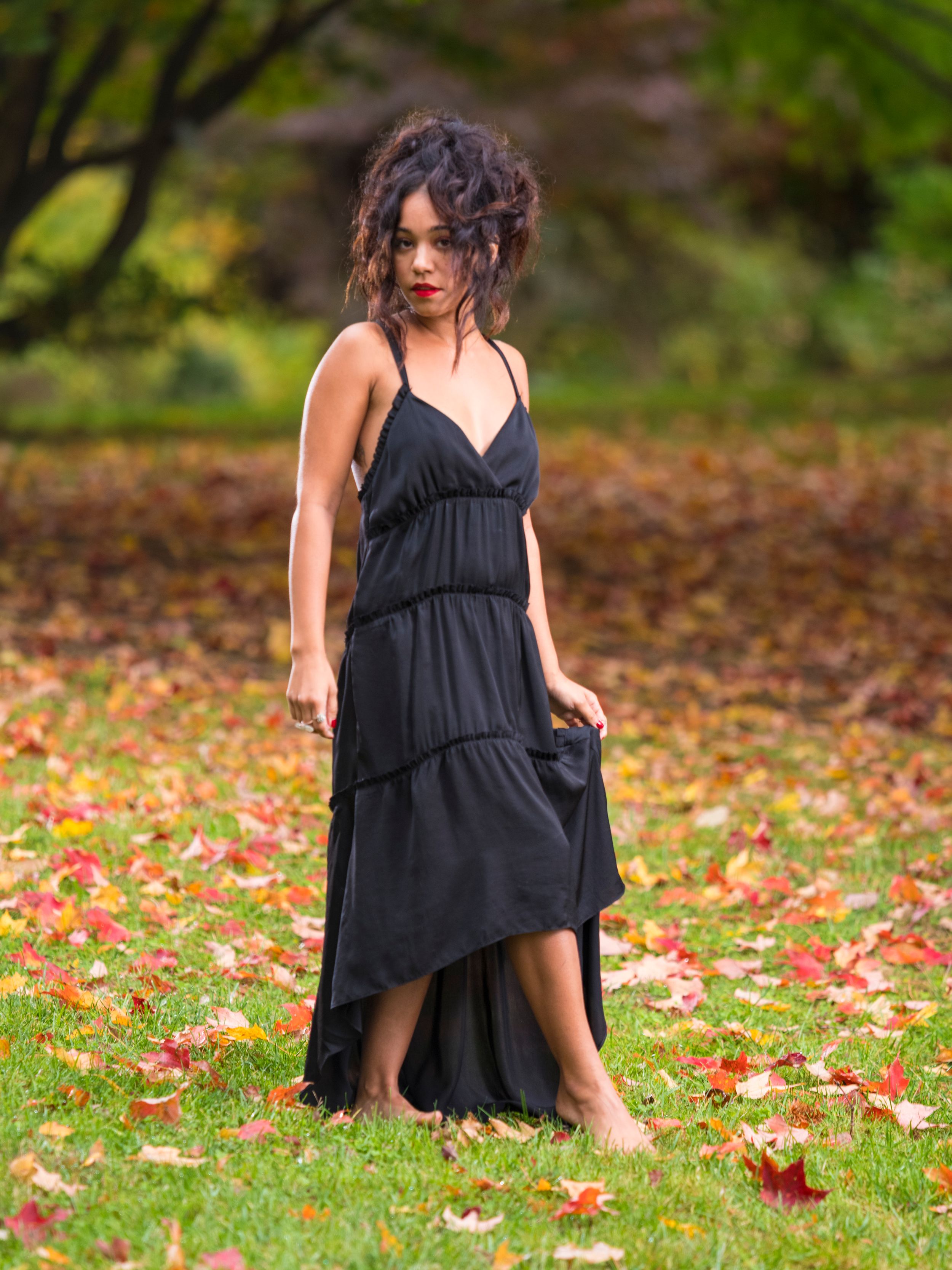 A person in a black tiered maxi dress stands among fallen autumn leaves in a garden setting.