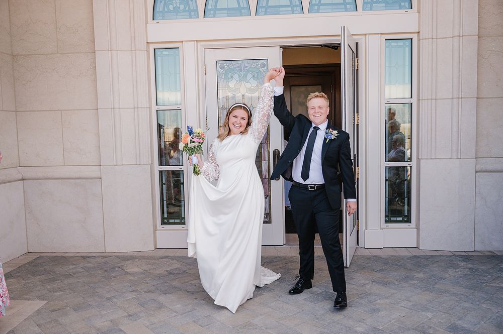 A happy couple celebrates outside a temple entrance with raised arms and joyful expressions.