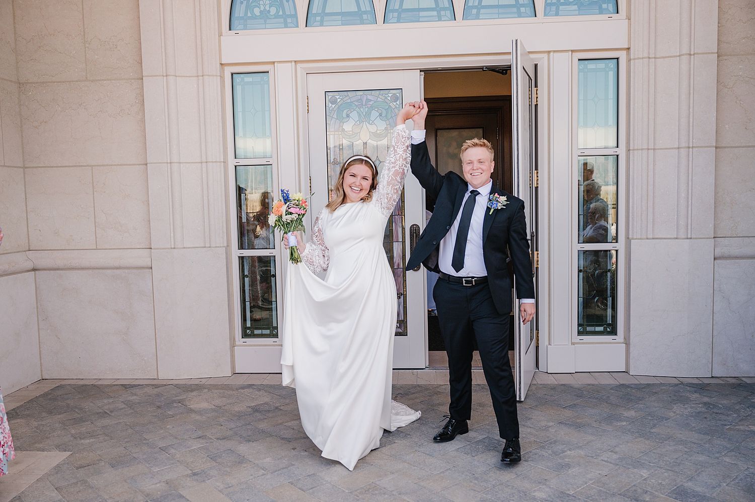 A happy couple celebrates outside a temple entrance with raised arms and joyful expressions.