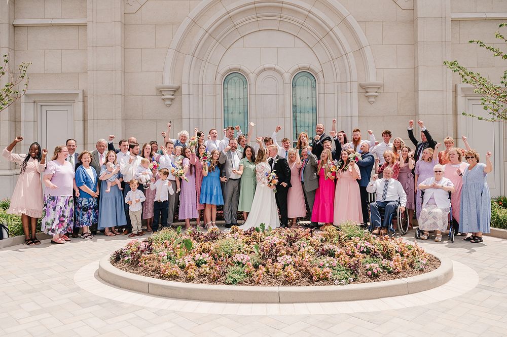 Large wedding party poses together outside ornate church building with arched windows and circular garden bed.
