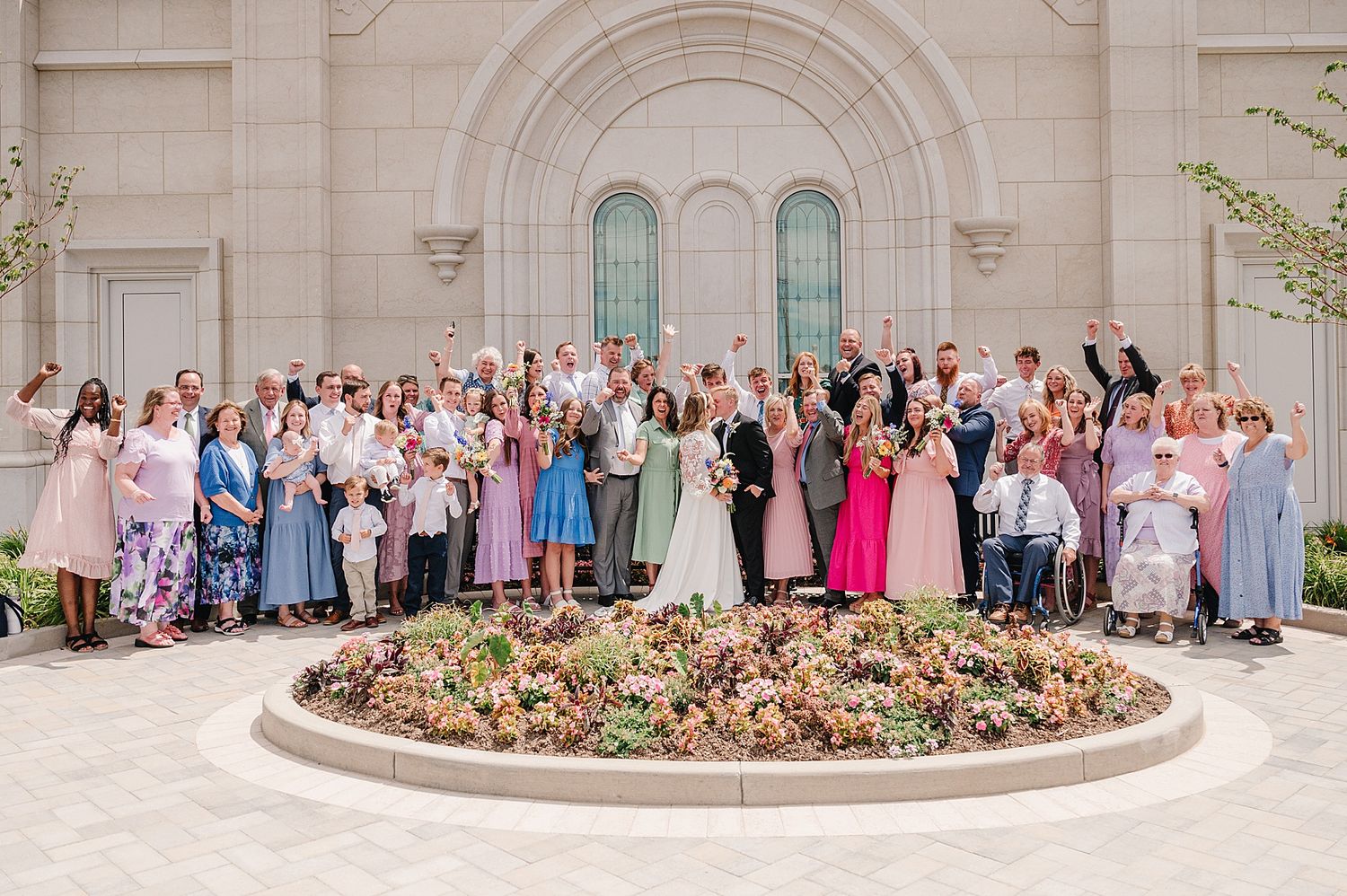 Large wedding party poses together outside ornate church building with arched windows and circular garden bed.