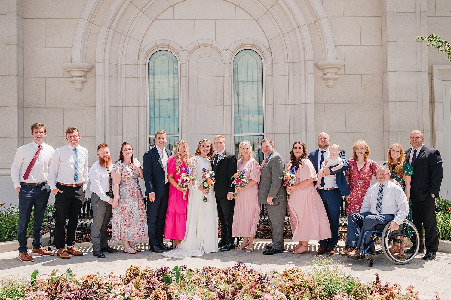 A large wedding party poses together in front of an elegant stone building with arched windows.