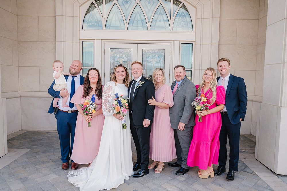 A wedding party poses outdoors in formal attire including pink bridesmaid dresses and suits in front of an arched doorway.