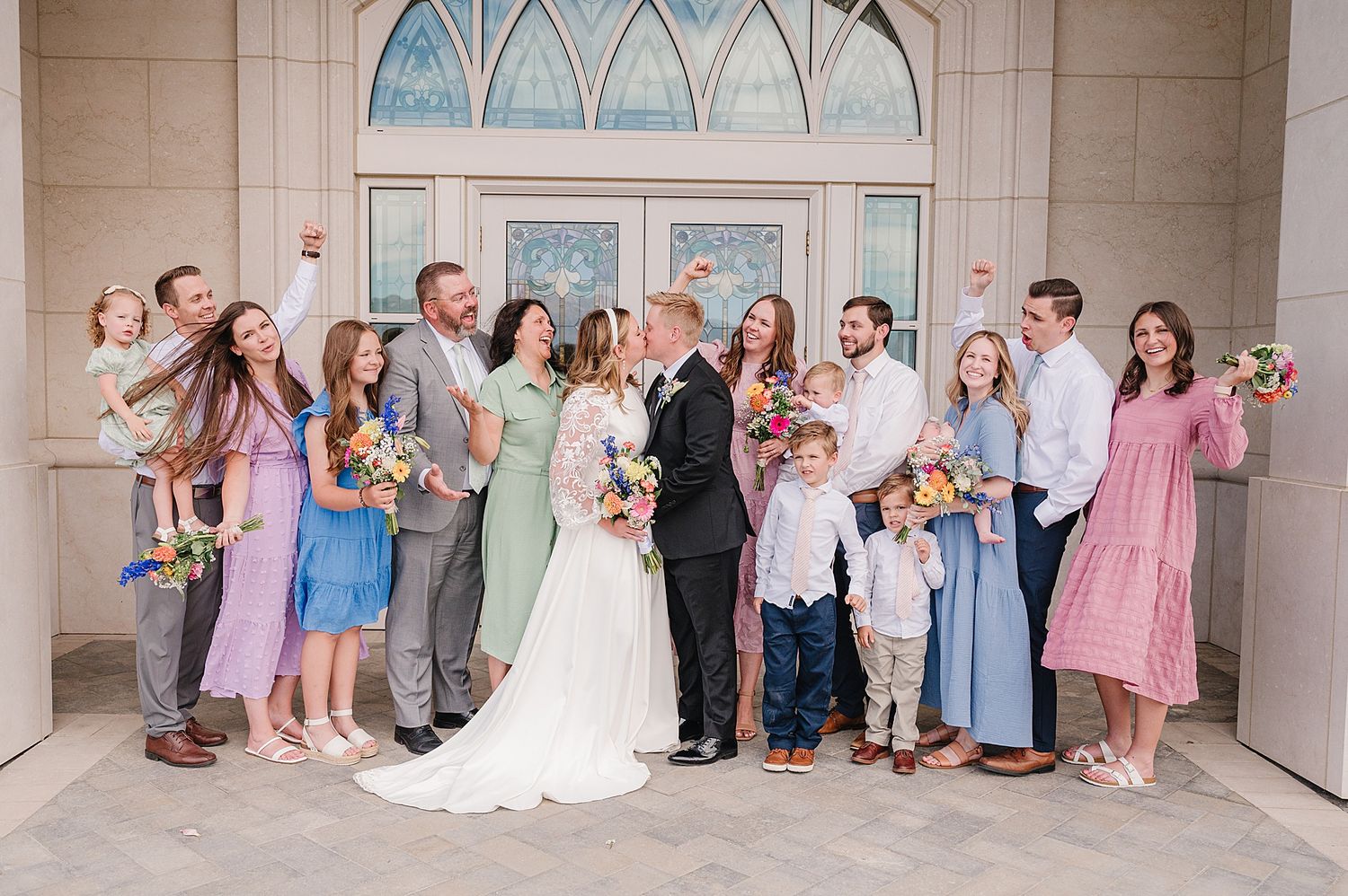 Wedding party in pastel dresses and suits poses together under an arched doorway of a cream-colored building.