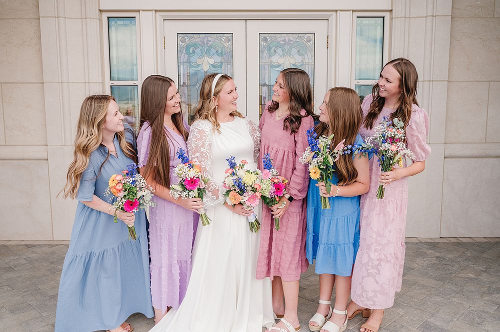 Bridesmaids in pastel dresses gather around bride in white dress for a candid moment outside church entrance.
