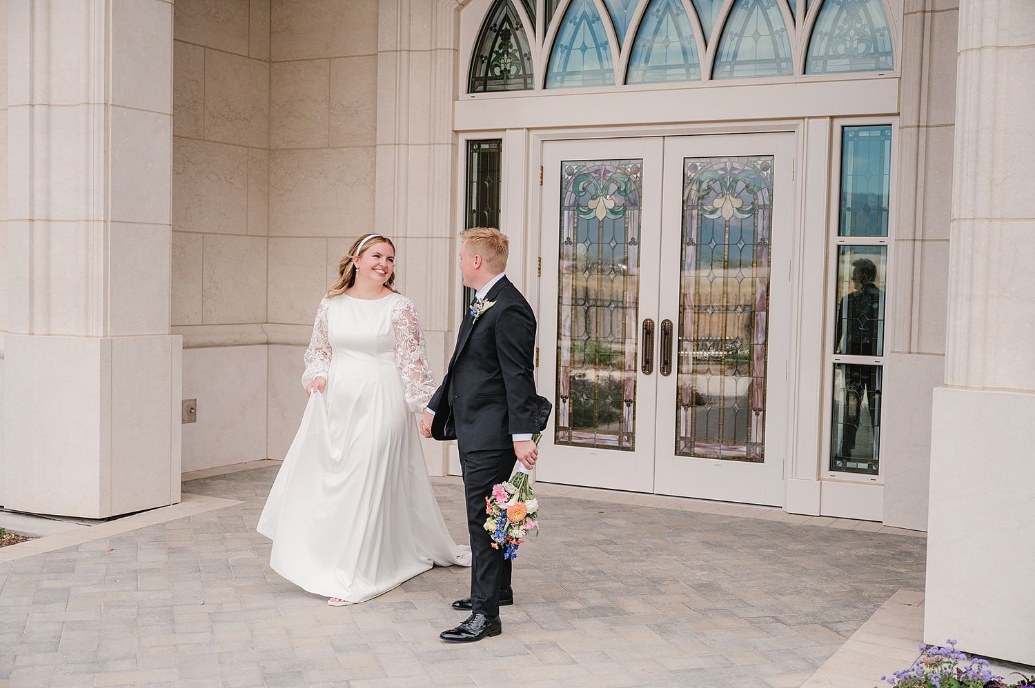 Two newlyweds share a joyful moment together outside an elegant building with arched windows and ornate doors.