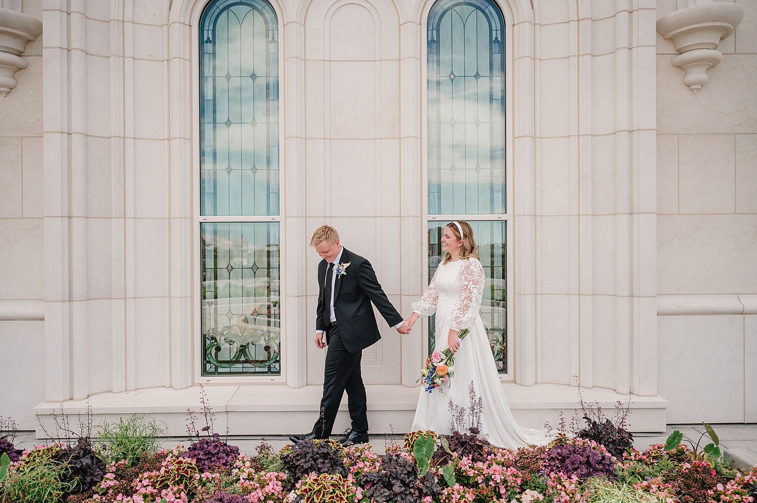 Couple poses against a light stone building with tall arched windows and colorful flower garden below.