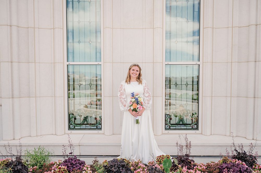 Bride in lace wedding dress holding colorful bouquet poses against church windows and flower garden.