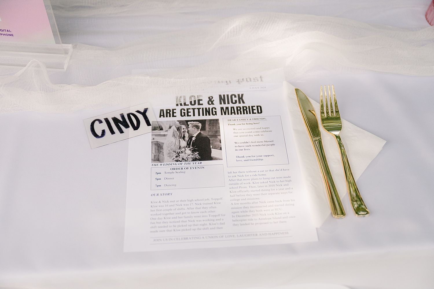 Wedding reception table setting with newspaper-style program and gold utensils on white tablecloth.