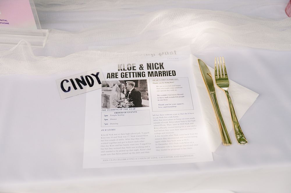 Wedding reception table setting with newspaper-style program and gold utensils on white tablecloth.