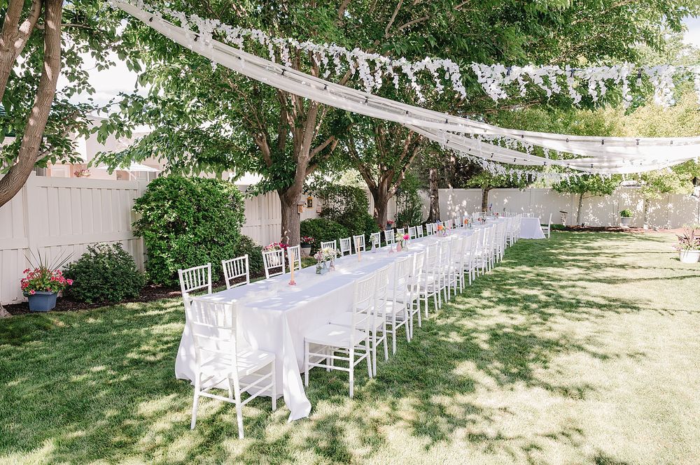 Elegant outdoor wedding reception setup with long white tables and chairs under string lights and tree canopy.