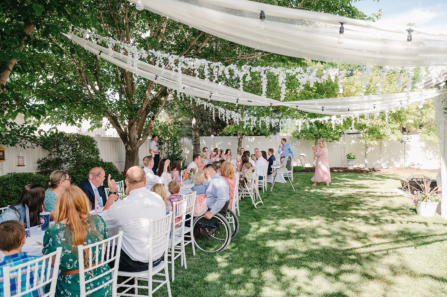 Outdoor wedding reception under white fabric canopy with guests seated at long tables on a sunny garden lawn.