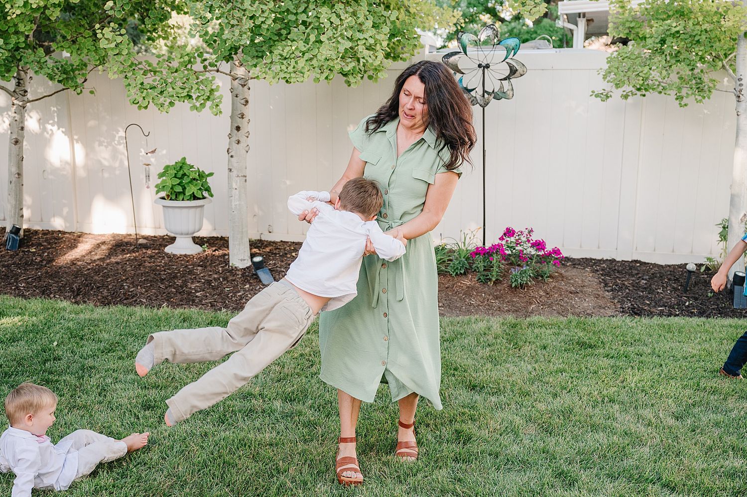 A person in a mint green dress plays with children on a grassy lawn near flowering plants and a white wall.