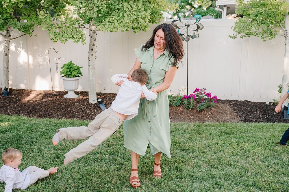 A person in a mint green dress plays with children on a grassy lawn near flowering plants and a white wall.