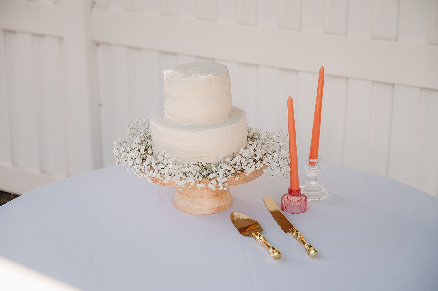 Simple white two-tier wedding cake adorned with baby's breath and coral accents on white tablecloth with gold utensils.