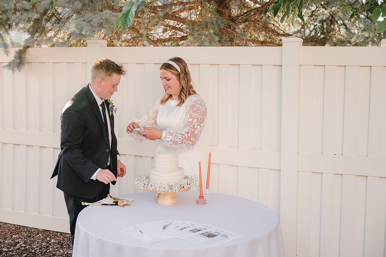 A couple slicing cake together at an elegant outdoor reception with white fence backdrop.