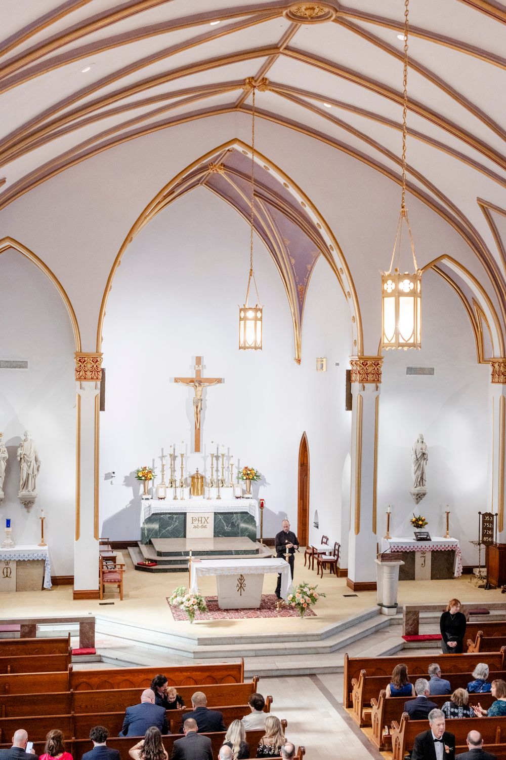 Interior of a Catholic church with gothic arched ceilings, hanging lanterns, and wooden pews during a ceremony.