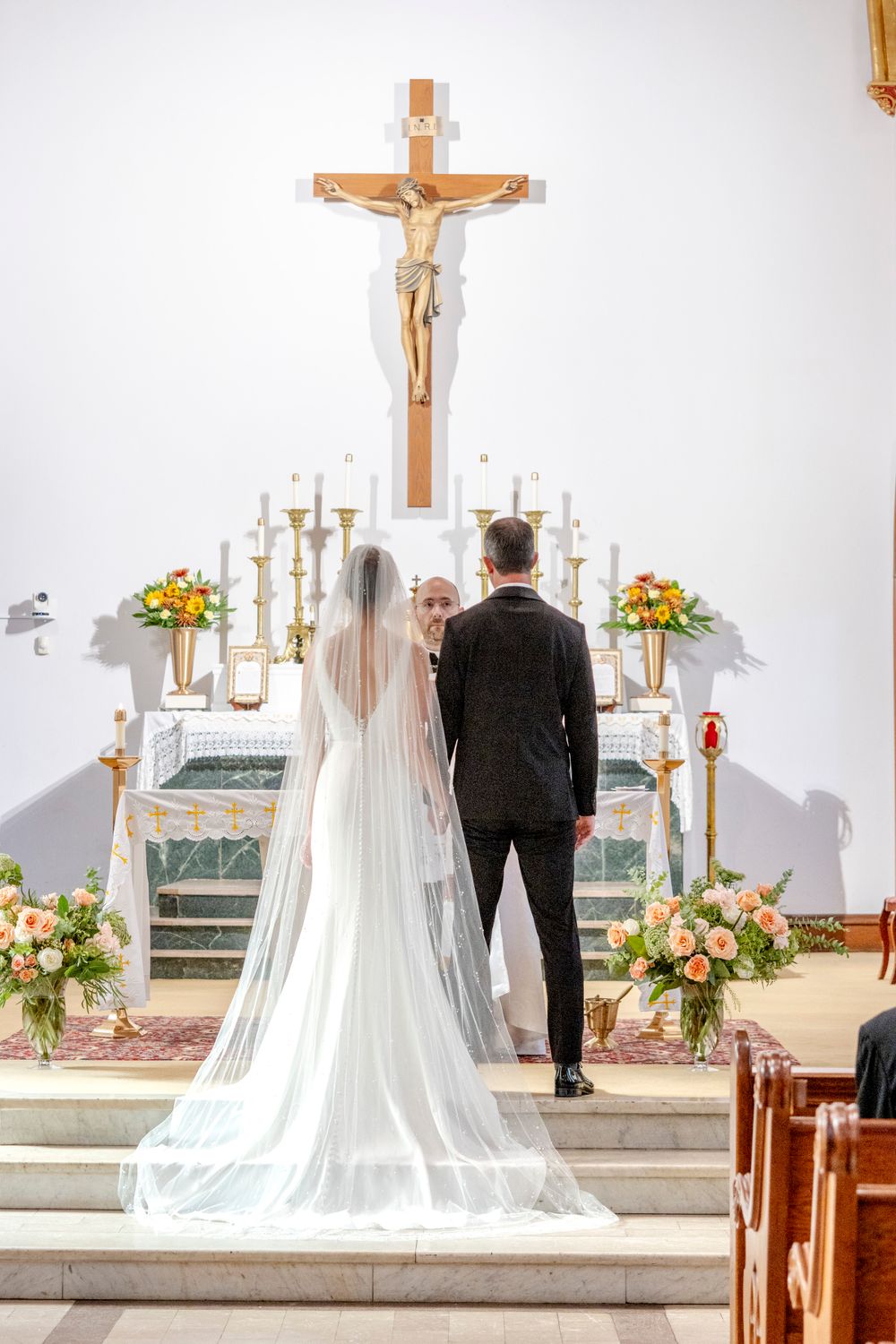 Wedding ceremony at church altar with crucifix, candles, and floral arrangements in soft lighting.