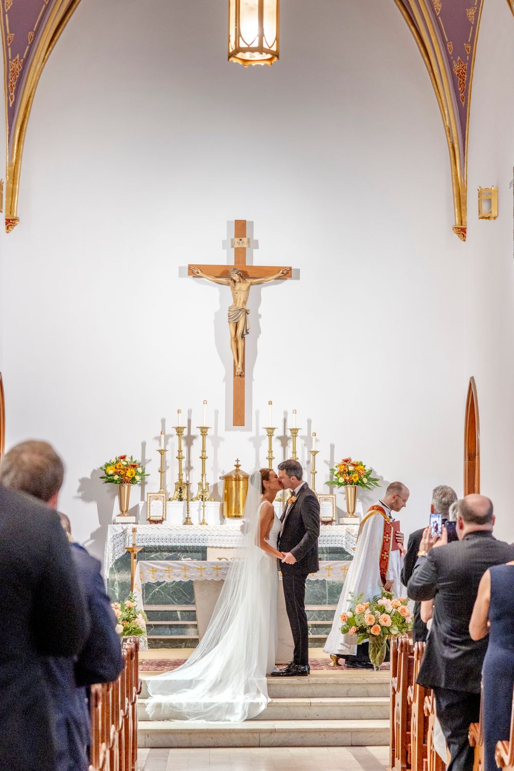 Wedding ceremony in progress beneath vaulted church ceiling with decorative gold trim and pendant lighting.