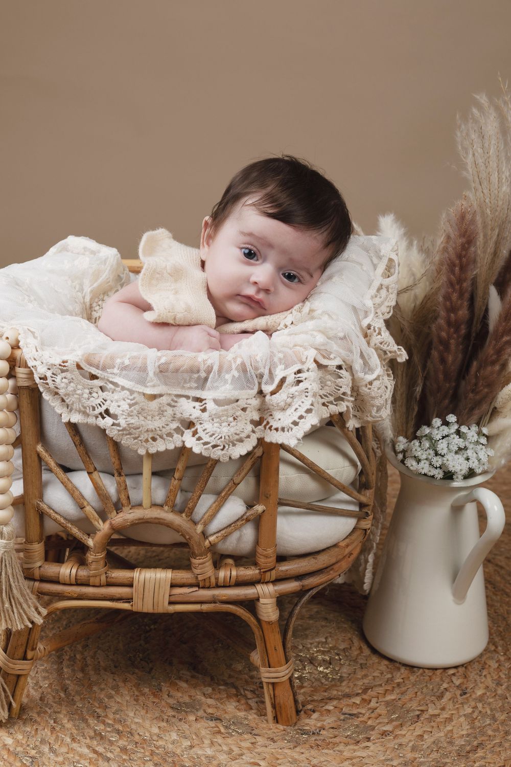 Newborn baby resting in a wicker bassinet with lace trim, surrounded by dried flowers in a boho nursery setting.