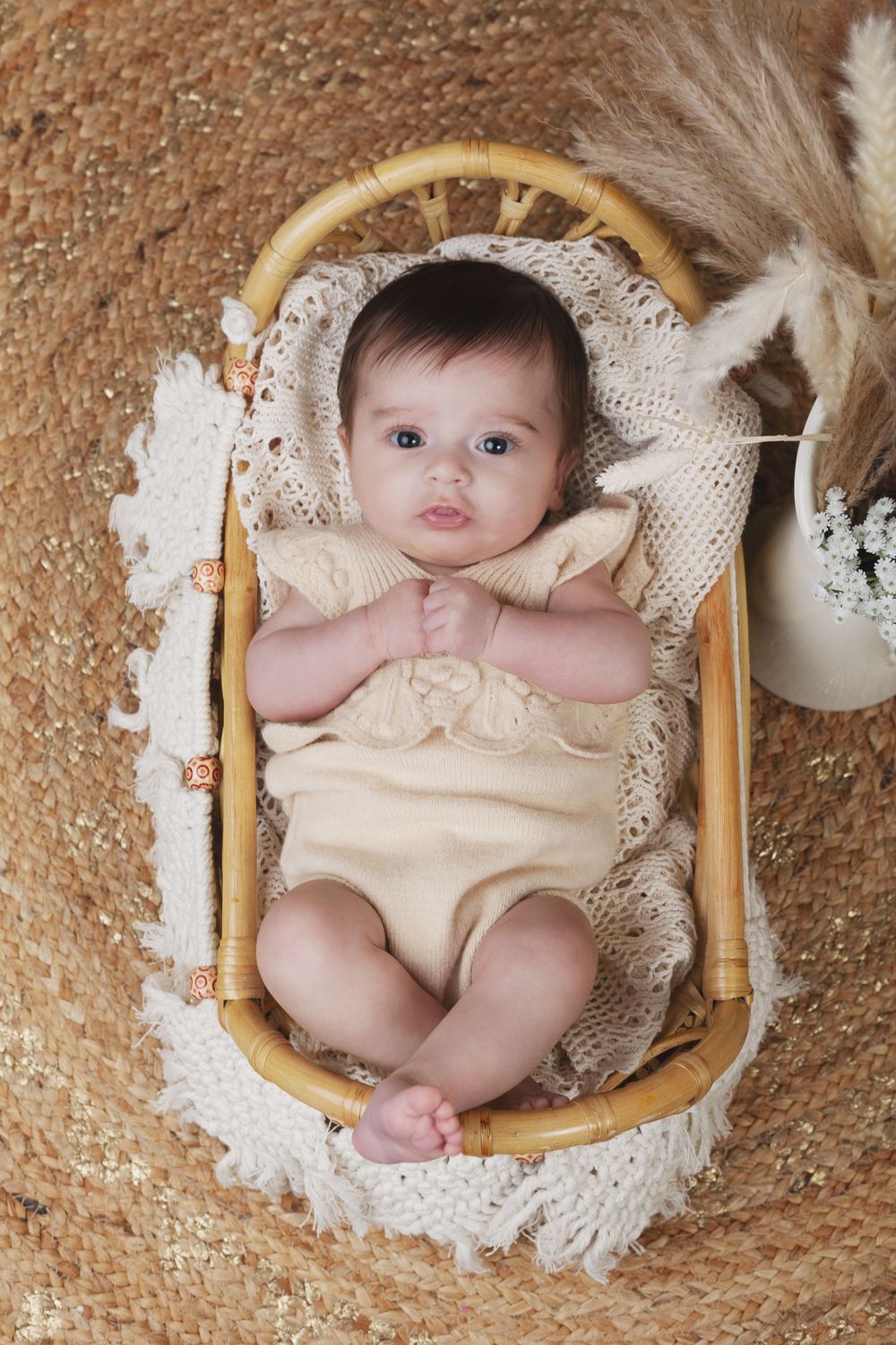 Adorable baby in yellow romper lying in a wicker Moses basket with crochet lining on a woven rug.