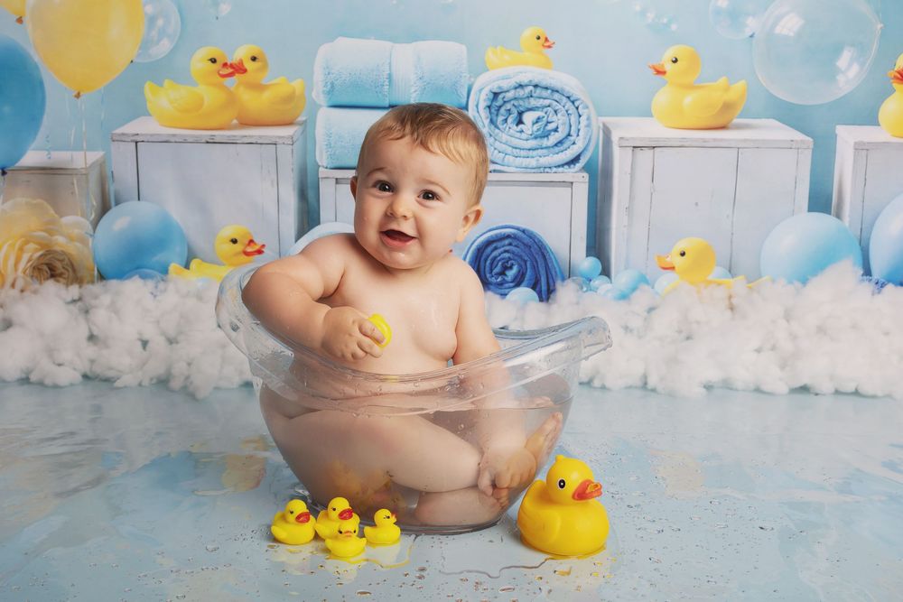 Baby sitting in bathtub with yellow rubber ducks and blue swirled decorations in background.