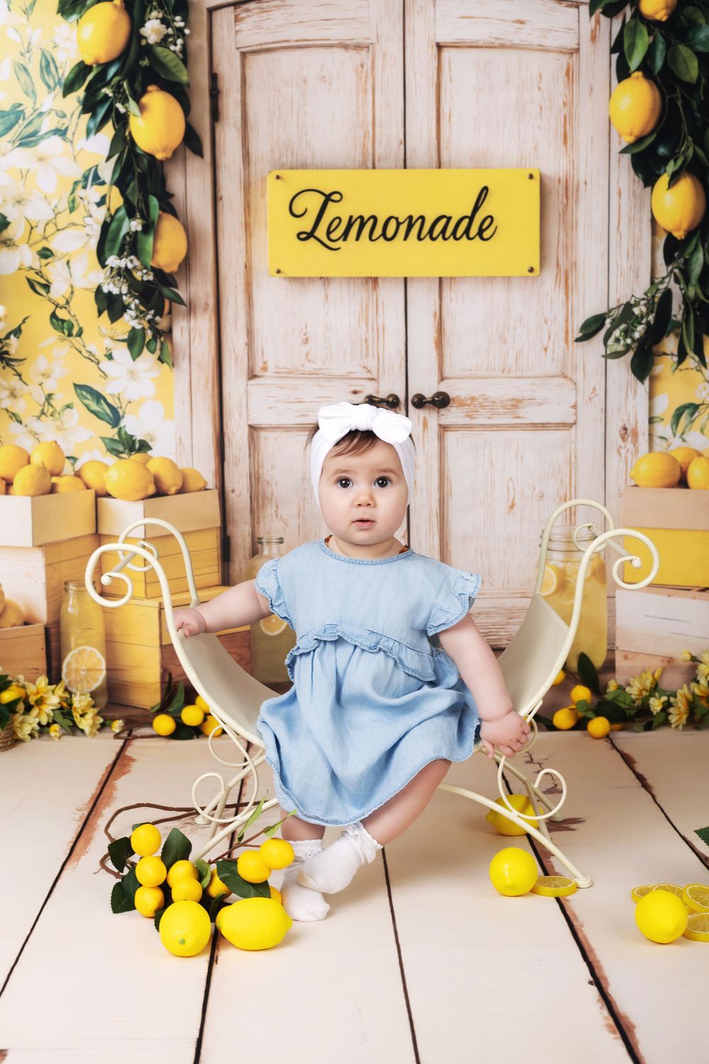 Adorable baby girl in blue dress sits among lemons before a rustic lemonade stand backdrop.