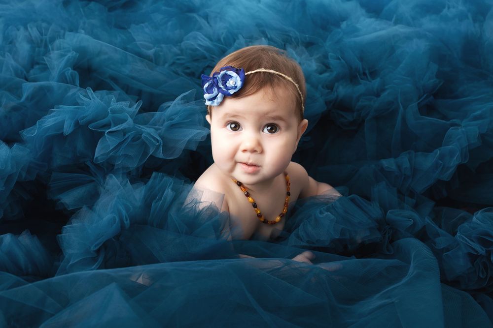 Adorable baby girl with blue flower headband surrounded by fluffy teal tulle fabric in a professional portrait photo.