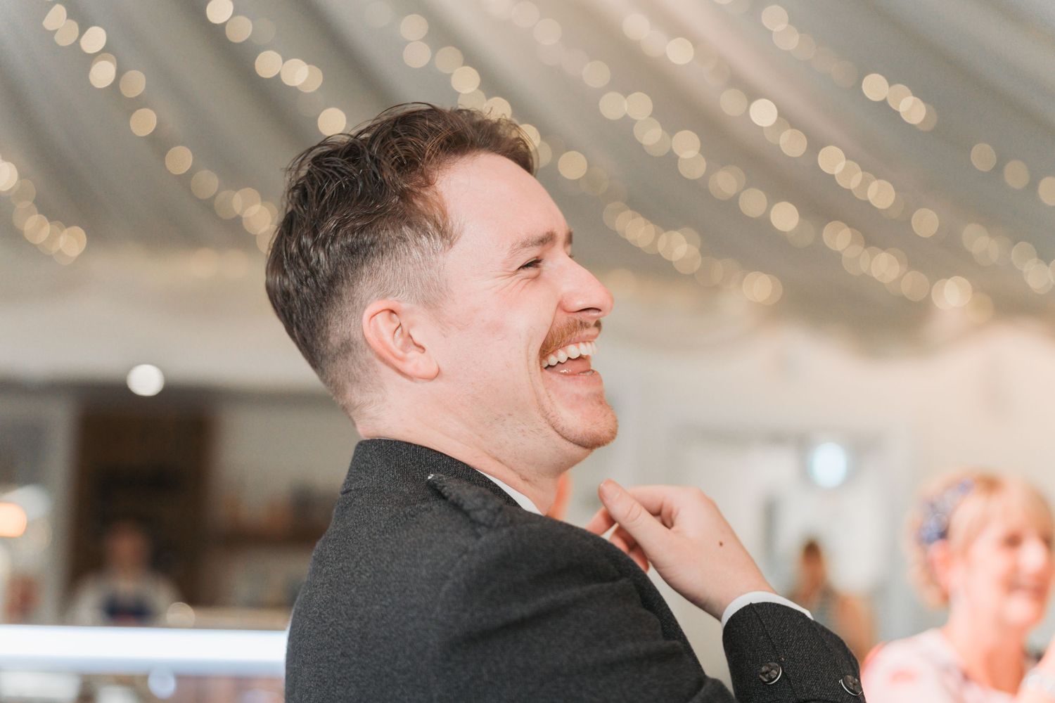 A person in a dark suit laughs joyfully at an indoor event with twinkle lights visible in the background.
