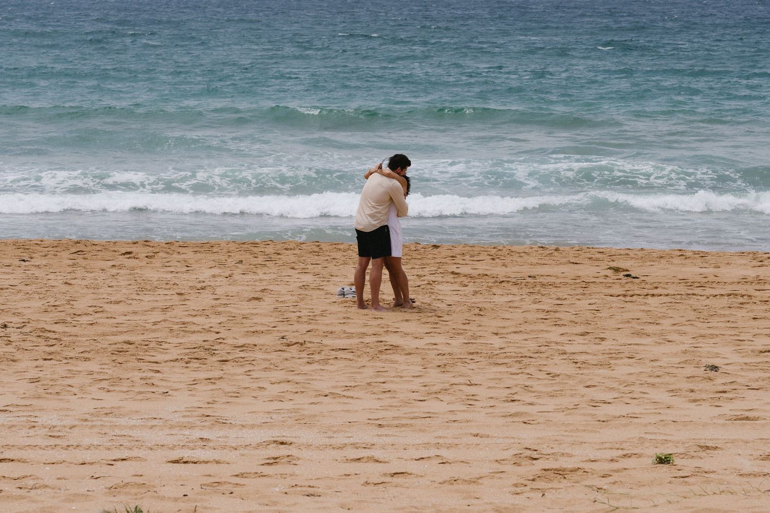 Two figures embrace on a sandy beach with ocean waves in the background.