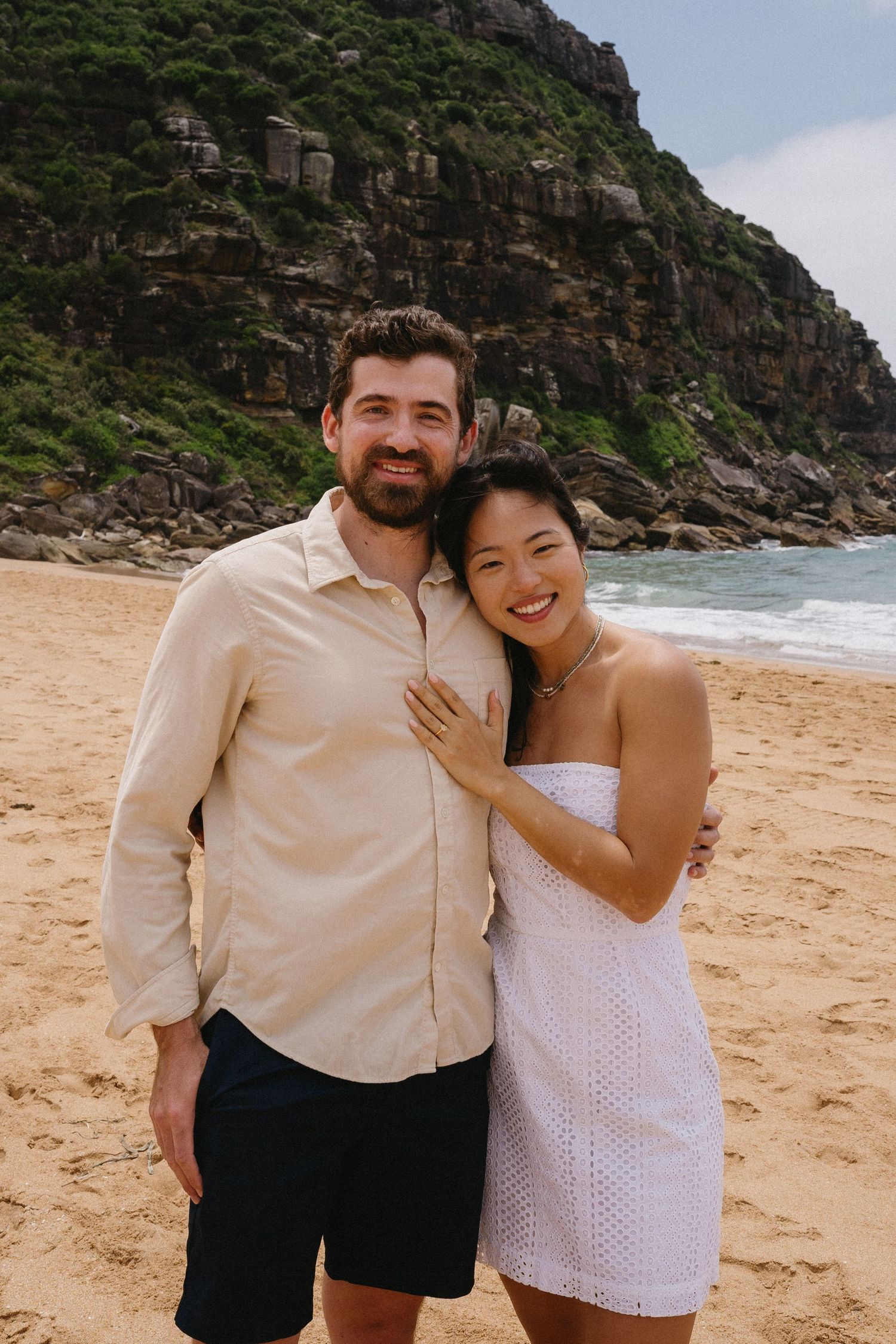 A happy couple posing on a beach with dramatic coastal cliffs in the background.