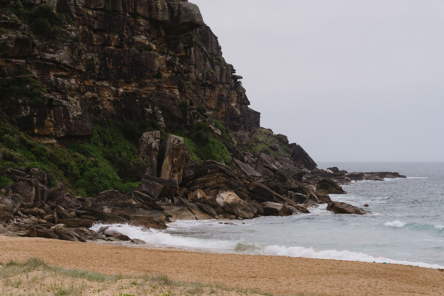Rocky coastal cliffs meet a sandy beach with crashing waves on a moody overcast day.