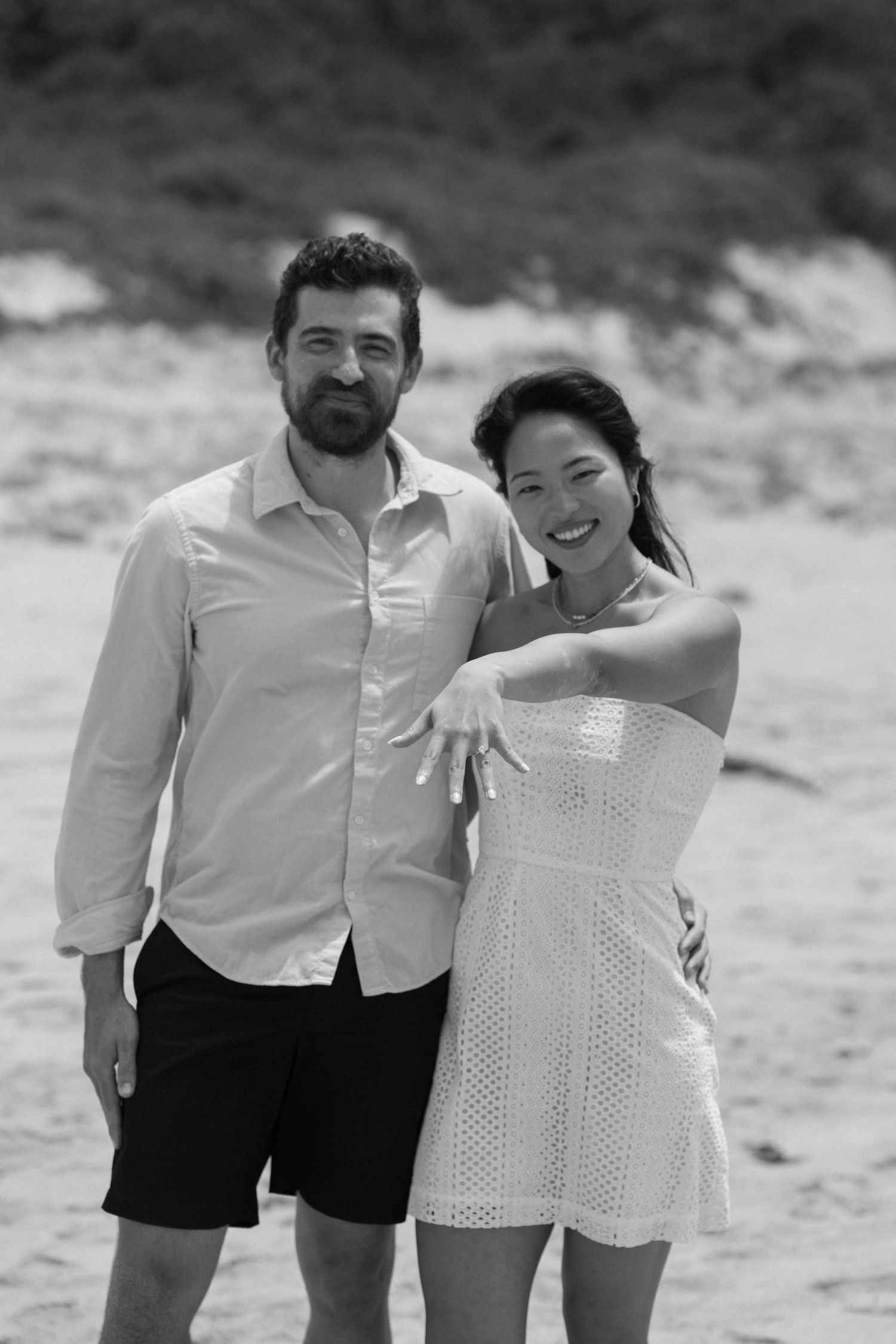A black and white portrait of a couple on the beach showing off an engagement ring.