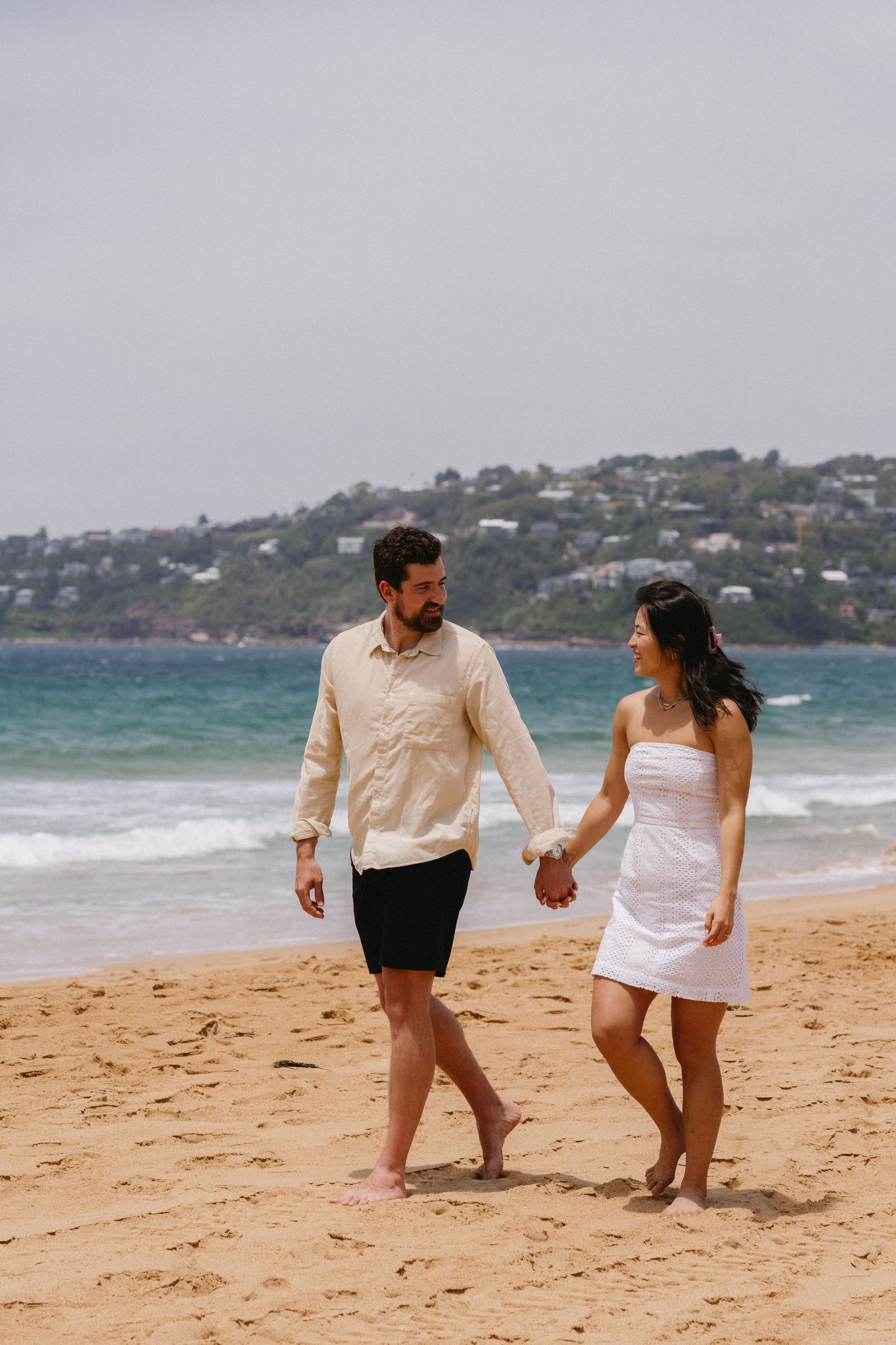 A couple walking hand in hand on a sandy beach with turquoise waters behind them.