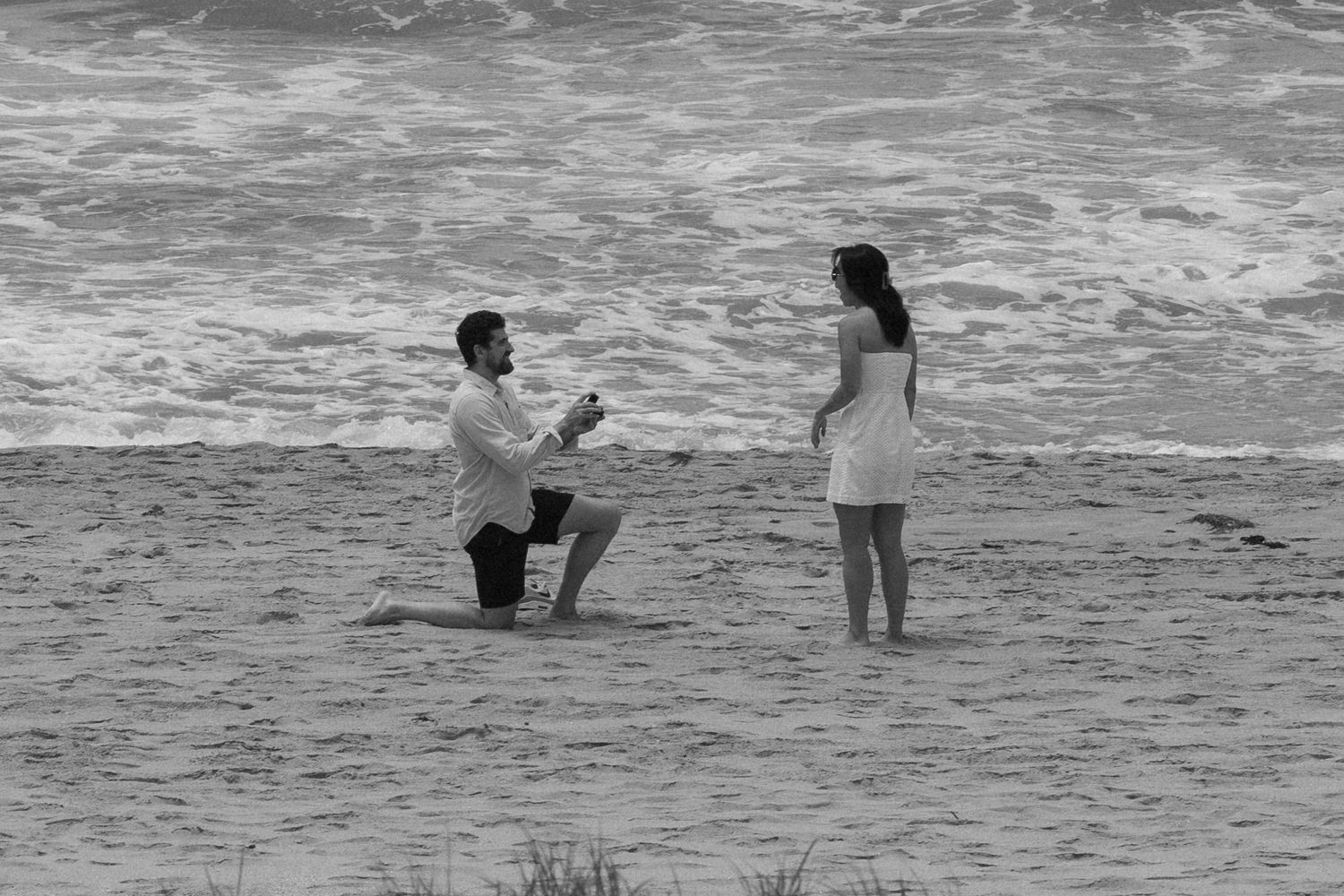 A black and white capture of a beach proposal with waves crashing in the background.