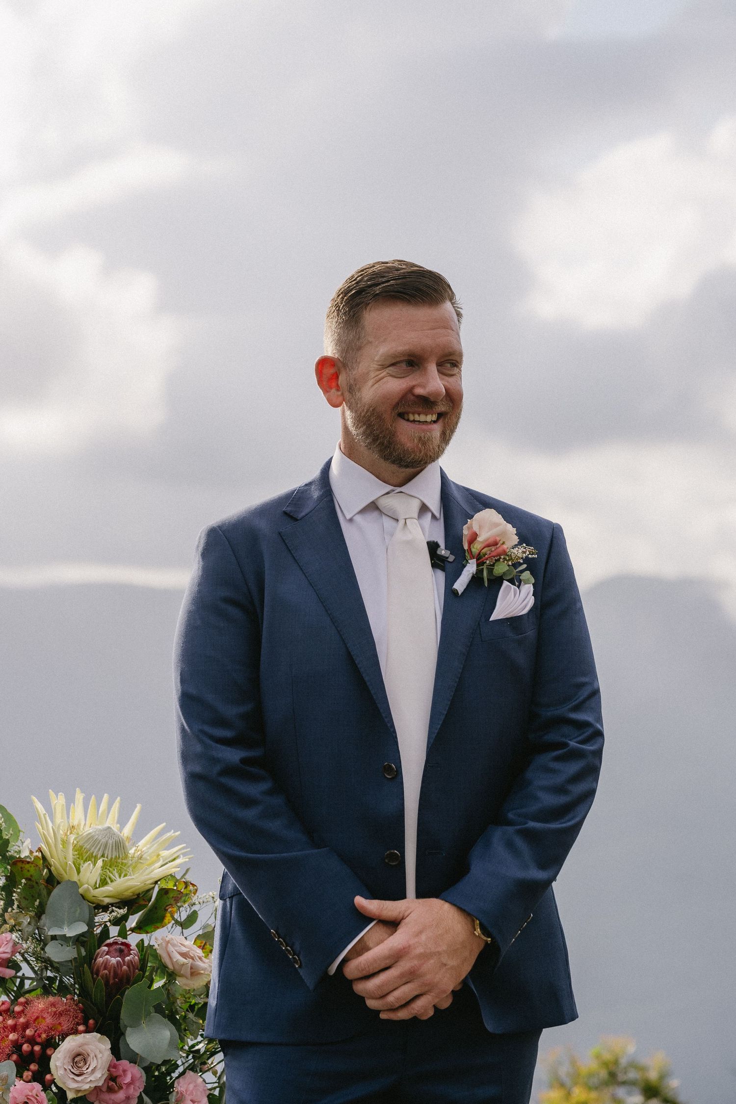Groom in navy suit with pink boutonniere smiles during outdoor mountain ceremony.