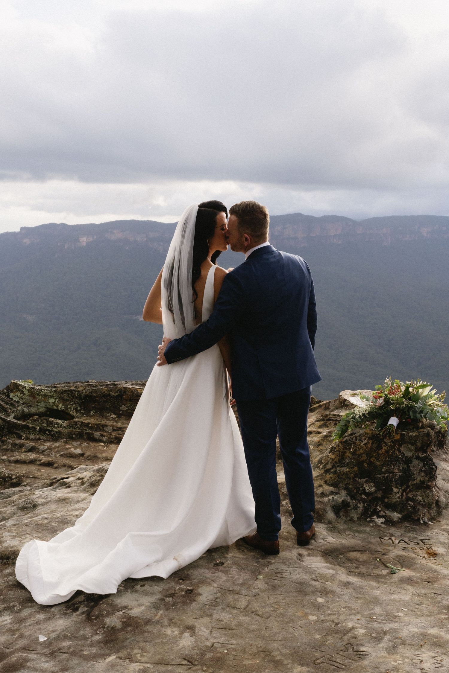 Romantic wedding couple embracing on mountain cliff with dramatic valley views.